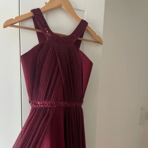 Vinröd långklänning med paljettdetaljer - Elegant vinröd långklänning med glittrande paljettdetaljer vid halsringning och midja. Klänningen har plisserad överdel och ett flytande, skirt tyg som ger en snygg siluett. Perfekt för bal eller festliga tillfällen.