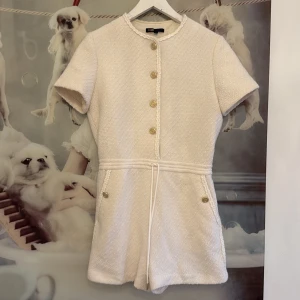 Ljus byxdress från Maje - Supersöt ljus från Maje med guldknappar framtill, korta ärmar och markerad midja med knyt. Klänningen har en strukturerad yta och dekorativa detaljer längs kanterna. Perfekt för dig som gillar en chic och stilren look. Strl. 38 men passform 34-36💖