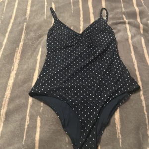 Svart prickig baddräkt med justerbara band - Supersnygg svart baddräkt med vita prickar och smala axelband med guldfärgade detaljer. Baddräkten har vadderade kupor och justerbara band för perfekt passform. Klassisk och tidlös modell som ger en stilren look på stranden.