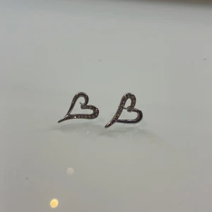 Silvriga hjärtformade örhängen - Säljer ett par stilrena örhängen i form av öppna hjärtan. De är silvriga och dekorerade med små glittrande stenar längs ena sidan av hjärtat. Perfekta för dig som gillar söta detaljer och vill ha något som sticker ut lite extra!!☺️