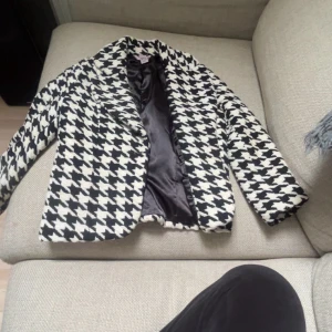 Svartvit rutig kavaj med mönster - Snygg svartvit kavaj med stort rutigt mönster, klassisk krage och helfodrad insida. Jackan har tre kvarts ärm och en rak passform som ger en cool och trendig look. Perfekt att slänga över en hoodie eller t-shirt för en extra edge. Säljer då jag köpte i fel storlek! Super trendig kappa