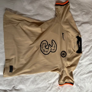 Chelsea FC beige bortatröja Nike - Chelsea FC:s officiella bortatröja i beige från Nike. Tröjan har korta ärmar med svart och orange detalj vid ärmslut, svart Nike-logga och klubbmärke på bröstet samt stor sponsorlogga framtill. Tillverkad i lätt och ventilerande Dri-FIT-material.