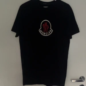 Svart Moncler t-shirt med logga helt ny   -    Helt ny svart moncler t shirt. Svart t-shirt från Moncler med stor vit och röd logga på bröstet. Klassisk passform och rund halsringning, perfekt för en clean och stilren look. Materialet känns mjukt och skönt mot huden.