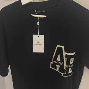 Svart t-shirt Anine Bing  - Svart t-shirt från Anine Bing med coolt broderat Letterman-märke i vitt och svart framtill. Klassisk passform med korta ärmar och rund hals. Perfekt statement-plagg för dig som gillar streetstyle och snygga detaljer. Nypris 1600