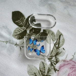 Transparent airpod fodral med blå fjärilar - Säljer ett transparent fodral med fina blå fjärilar på framsidan. Perfekt för att skydda dina hörlurar. Använt för airpods första generationen.  