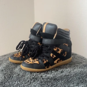 Leopardmönstrade svarta sneakers - Unika high top sneakers med leopardmönster i svart och beige, svarta mockadetaljer och svarta skinnpartier. Skorna har snörning, kardborreband och gummisula i beige. Perfekta för dig som vill sticka ut med din stil. Lite izabell marant stil