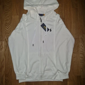 Vit hoodie från Polo Ralph Lauren  - Snygg vit hoodie från Polo Ralph Lauren i storlek M kan även passa S. Klassisk design med dragkedja, huva med snörning och broderad svart logga på bröstet. Mjuk bomullskänsla och ribbade muddar. Perfekt för en clean och stilren look.