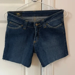 Mörkblå jeansshorts från Lee - Snygga mörkblå jeansshorts från Lee med råa kanter och klassisk femficksdesign. Shortsen har normal passform, gylf med knapp och dragkedja samt Lee-logga bak på linningen. Lågmidjade. Storlek är 27 i midjan men skulle uppskatta att den är mer som 26/25