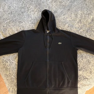 Svart hoodie från Lacoste med dragkedja - Svart hoodie från Lacoste med klassisk krokodil-logga på bröstet. Tröjan har huva med snörning, hel dragkedja och två stora fickor framtill. Tillverkad i mjuk bomull och har lång ärm. Perfekt för en chill och stilren look.