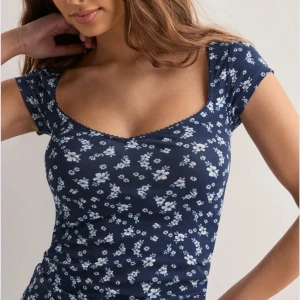 Blommig marinblå topp XS - Supersöt marinblå topp med ljusblå och vita blommor, korta ärmar och v-ringad hals med liten spetskant. Sitter figurnära och är gjord i mjuk bomullsmix. Perfekt för dig som gillar romantiska och trendiga plagg! Säljs för 200kr och helt oanvänd