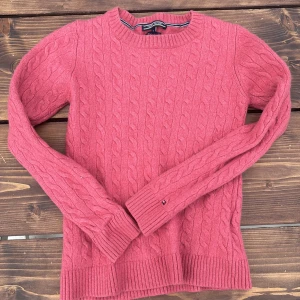 Rosa kabelstickad tröja Tommy Hilfiger - Kabelstickad rosa tröja från Tommy Hilfiger med rund halsringning och långa ärmar. Klassisk ribbad mudd vid ärmslut och nederkant samt diskret logga på ärmen. Perfekt för dig som gillar en stilren och tidlös look.