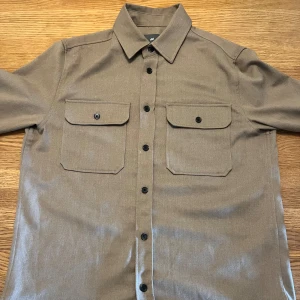 Beige overshirt från H&M med fickor - Stilren beige overshirt från H&M med klassisk krage och två stora bröstfickor med lock och knappar. Skjortan har långa ärmar och svarta knappar längs hela framsidan. Perfekt att bära öppen eller stängd för en avslappnad look. sitter som L
