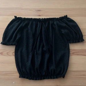 Svart topp LCW Casual XS - Supersöt svart topp från LCW Casual i storlek XS. Bär den som du vill, klassiskt eller off-shoulder. Perfekt för att matcha ditt humör och din stil! Toppen har puffiga korta ärmar, smockad struktur och resår både upptill och nertill för en snygg passform. Perfekt för dig som gillar trendiga och bekväma plagg med lite extra volym.
