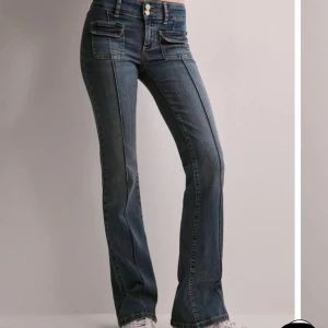 Blå pocket bootcut jeans - Snygga blå bootcut jeans med markerade sömmar framtill och dubbla fickor med lock. Jeansen har låg midja och två knappar i linningen. Materialet är klassisk denim med gul kontrastsöm, perfekt för en trendig look.💓💓