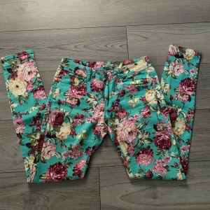 Blommiga jeans från C-in-c, strl 29 - Färgglada jeans från C-in-c i storlek 29 med ett stort blommönster i rosa, vinrött, gult och grönt på turkos botten. Perfekta för dig som vill sticka ut med en unik stil.