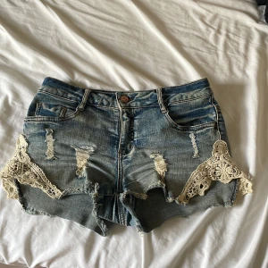 Rut&Circle jeansshorts med spetsdetaljer - Snygga blå jeansshorts från Rut&Circle med slitna detaljer och råa kanter. Utsmyckade med beige spets längs sidorna för en extra touch. Klassisk femficksmodell och knappstängning. Perfekta för sommarens alla äventyr!