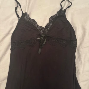 Svart spetsklänning med rosett - Svart kortklänning med tunna axelband och v-ringning. Klänningen har fina spetsdetaljer längs urringningen och under bysten samt en liten rosett framtill. Mjuk och lätt i materialet, perfekt för en chic och enkel look. Knappt använd. 