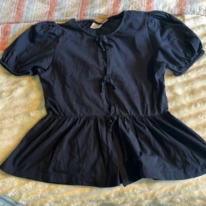 Svart blus med knyt från Gina Tricot - Supersöt svart blus från Gina Tricot med puffiga korta ärmar och tre knyt framtill. Blusen har en rund halsringning och en lätt utställd nederdel som ger en flowy känsla. Perfekt för dig som gillar en feminin och trendig look.