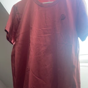 Röd Moncler t-shirt i bomull - Snygg röd t-shirt från Moncler i storlek L. T-shirten är i mjuk bomull, har rund halsringning och korta ärmar. På bröstet sitter Monclers klassiska logotyp i matchande röd ton. Perfekt för dig som gillar stilrena plagg med exklusiv känsla.