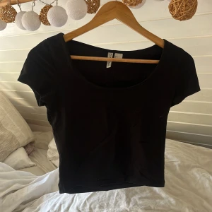 Svart basic topp från H&M Divided - En svart, enkel och tight topp från H&M Divided med rund halsringning och korta ärmar. Perfekt att matcha med jeans eller kjol för en clean look. Mjuk och stretchig känsla, passar dig som gillar minimalistisk stil.