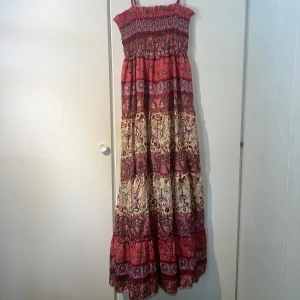 Lång boho-klänning, sommarklänning - Superfin långklänning i boho-stil med smock upptill och tunna axelband. Klänningen har ett färgstarkt mönster med fjärilar och blommor i vinrött, rosa och beige. Materialet känns lätt och flowy, perfekt för varma dagar. Volangdetaljer och mönstrade bårder ger en unik vibe. Somrig underbart skön! Passar xs-l