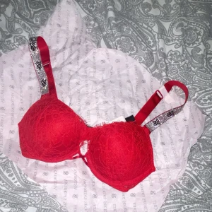 Röd spets-bh från Victoria's Secret - Snygg röd bh med spets från Victoria's Secret. Axelbanden är dekorerade med glittrande detaljer och små stenar för extra bling. Bh:n har vadderade kupor och en liten rosett framtill. Perfekt för dig som vill ha något extra fint under kläderna.