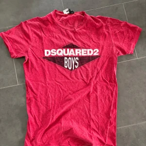 Röd Dsquared2 t-shirt med tryck - Riktigt snygg röd t-shirt från Dsquared2 med stort vitt och svart tryck på bröstet där det står 'DSQUARED2 BOYS'. Klassisk passform och rund hals. Perfekt för dig som vill sticka ut med ett coolt streetwear-plagg.