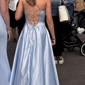 Ljusblå långklänning/balklänning med snörning - Säljer en elegant ljusblå långklänning i glansigt material med v-ringad omlottdesign framtill och öppen rygg med snygg snörning. Klänningen har smala axelband och ett svepande fall som ger en riktigt drömmig känsla.