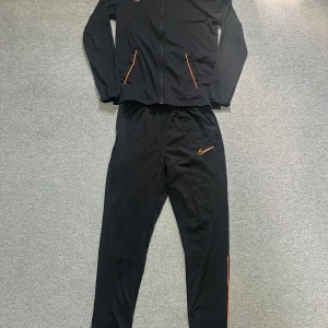 Nike svart träningsset Dri-Fit M - Nike svart träningsset i Dri-Fit material med orange detaljer och logga. Setet består av långärmad zip-jacka och matchande byxor med smal passform och orange ränder längs benen. Perfekt för dig som vill ha en sportig och clean look.