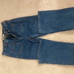 Blå raka jeans med hög midja - Klassiska blå jeans med raka ben och mid waist midja. Jeansen har fem fickor och stängs med dragkedja och knapp. Perfekta för en avslappnad och stilren look. Materialet är tåligt denim som håller formen hela dagen.