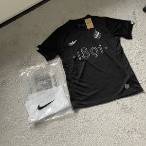 Svart AIK x Nike fotbollströja 1891 - Snygg svart AIK fotbollströja från Nike med trycket '1891' på bröstet, AIK:s klubbmärke och Nike-logga. Tröjan är kortärmad och tillverkad i ett lätt, sportigt material som andas. Perfekt för dig som vill visa ditt AIK-stöd både på och utanför planen.