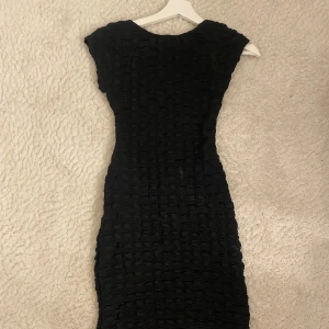 Svart kortklänning med struktur - Snygg svart kortklänning med coolt strukturerat tyg och figurnära passform. Klänningen har korta ärmar och en enkel, stilren design som passar perfekt när du vill sticka ut lite extra. Perfekt för dig som gillar minimalistisk men ändå unik stil.