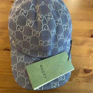 Blå keps från Gucci med GG-mönster - Snygg blå keps från Gucci med ikoniskt GG-mönster över hela kepsen. Justerbar rem baktill i mörkblått läder med guldfärgad detalj. Tillverkad i bomullsmix och har en klassisk böjd skärm. Perfekt för dig som vill sticka ut med en lyxig accessoar.