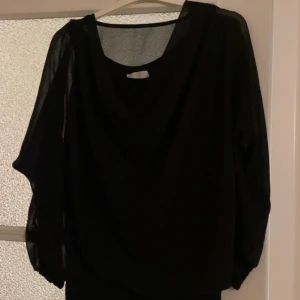 Svart blus med transparenta ärmar - Snygg svart blus med transparenta, lösa långärmar och vid passform. Blusen har en djupare urringning och är gjord i ett lätt, flowy material som ger en elegant känsla. Perfekt för dig som gillar stilrena och trendiga plagg.