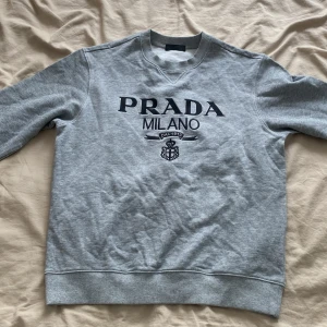 Prada Milano tröja - Snygg grå sweatshirt från Prada med svart logotyp och Milano-tryck framtill. Klassisk rund halsringning och långa ärmar. Tillverkad i mjuk polyester för en bekväm känsla. Perfekt för dig som gillar stilrena designer och vill ha något från ett lyxigt märke.