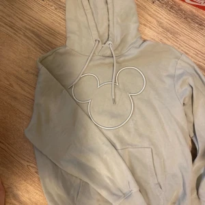 Beige Disney hoodie med Mickey Mouse - Snygg beige/grön hoodie från Disney med broderad kontur av Mickey Mouse på bröstet. Tröjan har huva med dragsko och en stor magficka. Perfekt för dig som älskar Disney och vill ha en chill och stilren look.