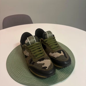 Valentino Rockrunner sneakers camouflage - Snygga Valentino Rockrunner sneakers i grön och beige camouflage med svarta detaljer. Skorna har snörning, platt sula och coola nitar på hälen. Materialmix av skinn, mocka och textil ger en lyxig känsla. Hör av dig vid fundering