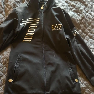 Svart EA7 Emporio Armani vindjacka S - Svart vindjacka från EA7 Emporio Armani med vita tryckdetaljer på bröstet och ärmen. Jackan har dragkedja framtill, två fickor med tryckknappar och en sportig look. Perfekt för dig som gillar stilrena och moderna plagg.