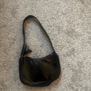 Svart axelväska i skinnimitation - Snygg svart axelväska med minimalistisk design och mjukt, glansigt material. Väskan har en enkel dragkedja upptill och ett brett axelband som gör den bekväm att bära. Perfekt för att få plats med det viktigaste när du är på språng.