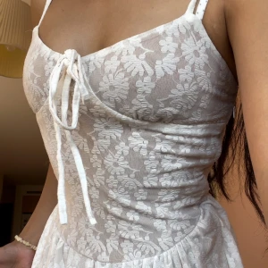 Vit lång spetsklänning med snörning - Romantisk långklänning i vit spets med blommigt mönster och smala axelband. Klänningen har v-ringning med snörningsdetalj framtill och en lätt utställd kjol. Perfekt för sommarens alla tillfällen och ger en dreamy vibe.
