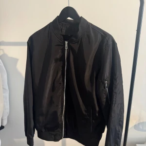 Svart bomberjacka med dragkedja - Snygg svart bomberjacka med klassisk passform och silvrig dragkedja framtill. Jackan har ribbade muddar vid ärmslut och midja samt en extra ficka med dragkedja på ena ärmen. Perfekt för dig som gillar en clean och stilren look.