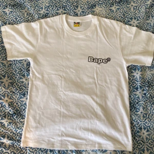 Vit graphic Bape T-shirt - Vit Bape T-shirt med klassisk logga på bröstet och stort svartvitt tryck med olika Bape-grafiker på ryggen. Normal passform och tillverkad i 100% bomull. Säljer den eftersom den är för stor för mig, HELT oanvänd. Ifall måtts söks fråga i dm’s. Priset är INTE fast