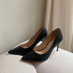 Svarta pumps från Guess med spetsig tå - Klassiska svarta pumps från Guess i skinn med elegant spetsig tå och hög klack. Snyggt minimalistisk design med diskret G-detalj i silver på hälen. Perfekta för dig som vill ha en stilren och tidlös look.