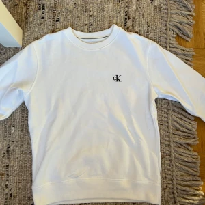Vit sweatshirt från Calvin Klein - Vit sweatshirt från Calvin Klein med diskret CK-logga broderad på bröstet. Klassisk rund halsringning och långa ärmar. Mjuk och skön i bomullsmaterial, perfekt för en clean och stilren look.