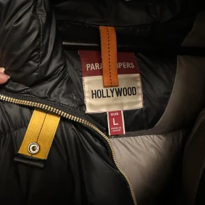 Svart dunjacka Parajumpers L - Säljer en svart dunjacka från Parajumpers, modell Hollywood. Jackan har snygga detaljer med orange och gula inslag, patch på ärmen och dragkedja framtill. Fodret är grått och materialet känns slitstarkt och varmt, perfekt för kalla dagar. Priset går att diskutera