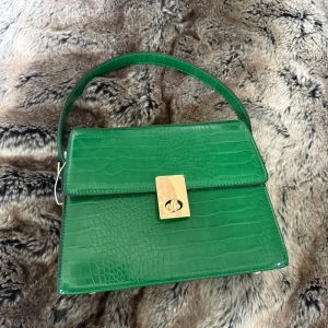 Grön handväska från Laphrodite Paris - Stilren grön handväska från Laphrodite Paris i croco-mönstrat syntetmaterial. Väskan har guldfärgat lås framtill, ett stadigt handtag och flera fack med dragkedja både inuti och på baksidan. Aldrig använd. Nypris 1000 kr.