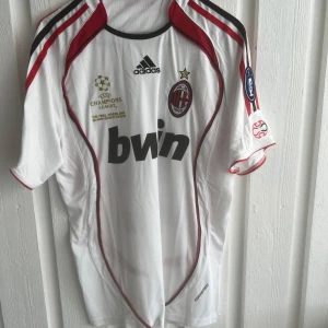 AC Milan Kaka Champions League tröja - AC Milan fotbollströja med Kaka och nummer 22 på ryggen. Vit med röda och svarta detaljer, Champions League-märken på ärmarna och Bwin som sponsor. Adidas-logga och klubbmärke på bröstet. Tillverkad i lätt funktionsmaterial, kortärmad modell.