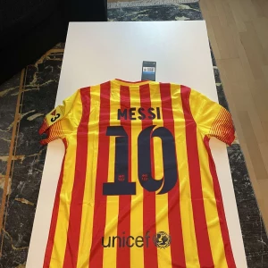 Barcelona Messi fotbollströja randig - Säljer en officiell FC Barcelona fotbollströja med Messi 10 på ryggen. Tröjan är kortärmad, har röda och gula vertikala ränder, klubbmärke på bröstet och Qatar Airways-tryck. Tillverkad i lätt och ventilerande Dri-FIT-material. Perfekt för dig som älskar Barca och Messi.