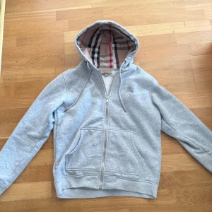 Ljusgrå hoodie från Burberry - Snygg ljusgrå hoodie från Burberry med klassiskt rutigt foder i huvan. Tröjan har dragkedja framtill, snörning i huvan och två stora fickor. Perfekt för dig som gillar stilrena och bekväma plagg med en touch av lyx.