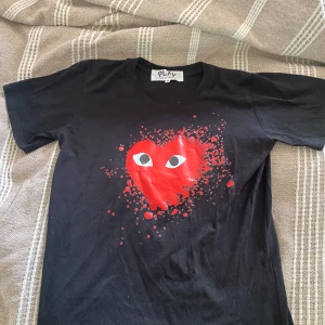 Svart Comme des Garçons Play t-shirt - Svart t-shirt från Comme des Garçons Play med ikoniskt rött hjärta med ögon på bröstet. Klassisk passform och rund hals. Trycket har en cool splash-effekt som gör den extra unik. Perfekt för dig som vill sticka ut med streetstyle.